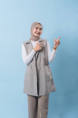 Gri yelek ve beyaz gömlek giyen bir kadın önemli bir şeyi işaret ediyor. Yüzünde bir gülümseme var ve bir türban takıyor.