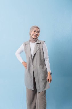 Gri yelek ve beyaz gömlek giyen bir kadın kameraya gülümsüyor. Başörtüsü takıyor ve kollarını kavuşturuyor.