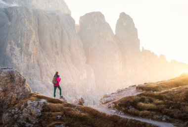 Gün batımında dağ yolunda sırt çantalı genç bir kadın ve dağ kanyonu. Dolomitler, İtalya 'da sonbahar. Renkli manzara, yolda kız, kayalar, sonbaharda altın güneş ışığı. Gezinti ve yürüyüş