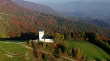 Tepedeki küçük kilisenin havadan görünüşü ve sonbaharda gündoğumunda alçak bulutlar. Slovenya. Dağdaki güzel şapelin insansız hava aracı manzarası sisli yeşil çayırlar, ağaçlar, sonbaharda şafak vakti gökyüzü. Peyzaj