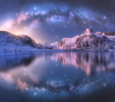 Samanyolu, deniz kıyısının üzerindeki kemer ve kışın kış mevsiminde karla kaplı dağlar. Lofoten Adaları, Norveç. Kutup manzarası mavi yıldızlı gökyüzü, kavisli samanyolu suya, karlı kayalara yansıyor. Boşluk