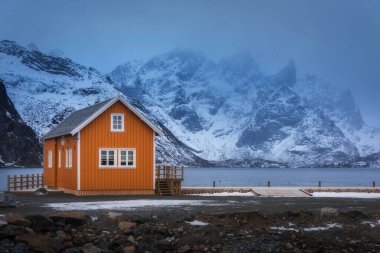 Deniz kıyısında turuncu rorbu ve alacakaranlıkta sisle kaplı dağ. Lofoten Adaları, Norveç. Geleneksel Norveç kuyusu, alacakaranlıkta karlı kayalar ile kasvetli kış manzarası. Eski balıkçıların evi