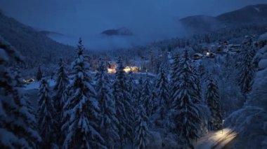 Aerial view of fairy forest in snow, frozen lake and house with lights at night in winter. Top view from drone of mountain village, pond, illumination, snowy pine trees, low clouds at dusk in Slovenia