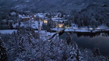 Aerial view of beautiful Bohinj lake in Slovenia at night in winter. Top view from drone of snowy mountains, trees, church, bridge, road, city lights, houses, reflection in water, purple sky at sunset