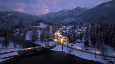 Aerial view of beautiful Bohinj lake in Slovenia at night in winter. Top view from drone of snowy mountains, trees, bridge, cars on road, city lights, houses, reflection in water, purple sky at sunset