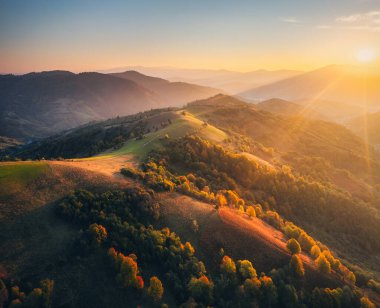 Aerial view of beautiful hills and mountains in fog at sunset in autumn in Ukraine. Colorful landscape with foggy woods, meadows, golden sunlight. Orange trees. Forest in fall. Drone view. Nature