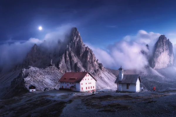 Mountains in fog with beautiful house and church at night in summer. Landscape with high rocks, blue sky with moon. Rocky mountain peaks in clouds. Tre Cime in Dolomites, Italy. Alps at twilight