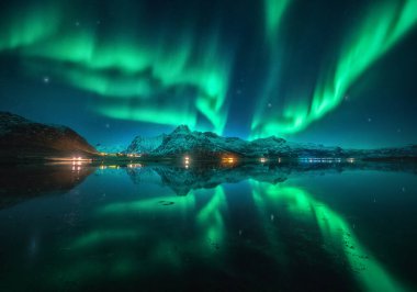 Kuzey ışıkları karlı dağların üzerinde, deniz, gece vakti Lofoten, Norveç 'te sudaki yansıma. Aurora borealis ve karla kaplı kayalar. Kutup ışıklarıyla kış manzarası, şehir ışıkları, yıldızlarla gökyüzü