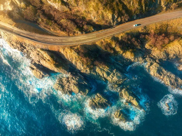Aerial view of road, rocky sea coast with waves and stones at sunset in Lofoten Islands, Norway. Landscape with beautiful road, transparent blue water, rocks. Top view from drone of highway in summer