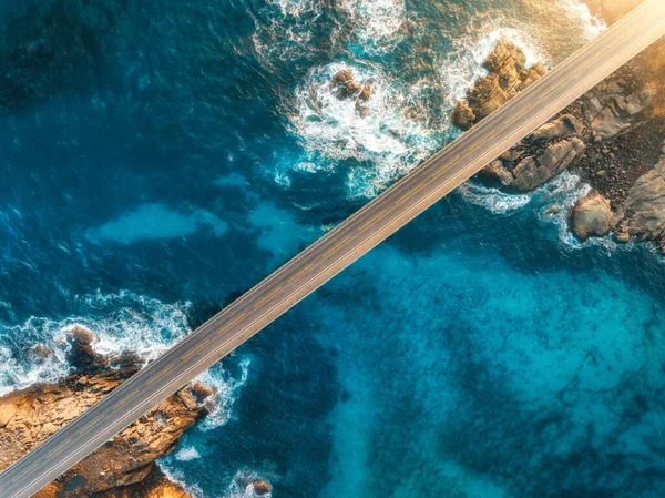 Aerial view of bridge, sea with waves and stones at sunset in Lofoten Islands, Norway. Landscape with beautiful road, transparent blue water, rocks. Top view from drone of highway in summer. Transport