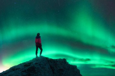 Kuzey ışıkları ve gece dağın zirvesinde genç bir kadın. Aurora Borealis ve kayanın tepesindeki yalnız kız silueti. Kutup ışıklarıyla manzara. Parlak aurora ile yıldızlı gökyüzü. Seyahat arkaplanı