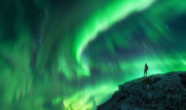 Kuzey ışıkları ve gece dağın zirvesinde genç bir kadın. Aurora Borealis ve kayanın tepesindeki yalnız kız silueti. Kutup ışıklarıyla manzara. Parlak aurora ile yıldızlı gökyüzü. Seyahat arkaplanı