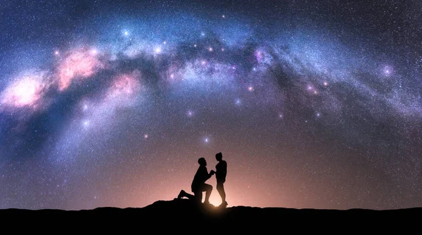 Milky Way with silhouettes of a man making marriage proposal to his girlfriend and starry sky at night. Silhouette of lovers. Couple, relationship. Milky way with people. Universe. Space background
