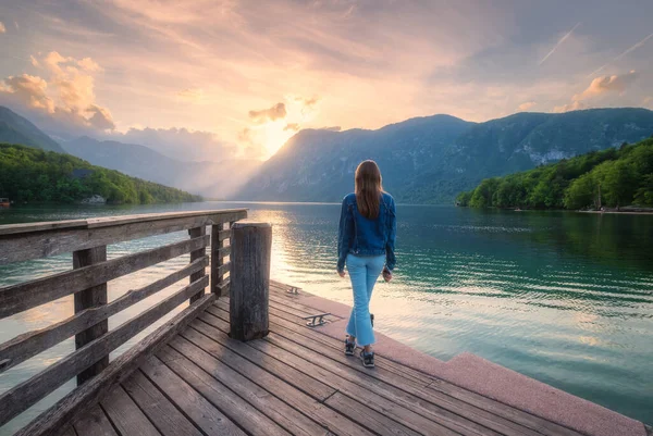 Ahşap iskelede, dağlarda ve baharda gün batımında Bohinj gölünde bir kadın. Kızım, suda yansıyan göl, dağlarda yeşil orman ve yazın renkli gökyüzü. Slovenya 'daki Triglav Ulusal Parkı