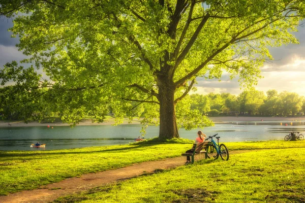 Woman sitting on bench and mountain bike, green trees and grass, lake at sunset in spring. Colorful landscape with resting girl, bicycle, river in park in summer. Sport and travel. Biking. Nature