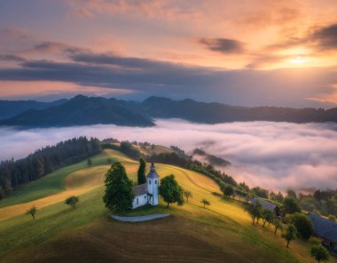 Slovenya 'da yaz aylarında sisli bir gündoğumunda tepedeki küçük kilisenin pembe alçak bulutlar üzerindeki görüntüsü. Dağdaki güzel şapelin sisli manzarası, yeşil çayırlar, ağaçlar, ilkbaharda şafak vakti turuncu gökyüzü.