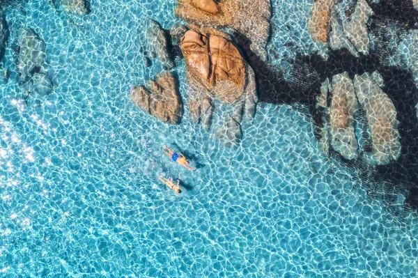 Mavi denizde yüzen adam ve kadının havadan görünüşü, kayalık plaj, yazın günbatımında, İtalya, Sardunya 'da. Tropikal arka plan. Şeffaf gök mavisi su, insanlar ve taşlar. Üst dron görüntüsü. Aşağı görünüm