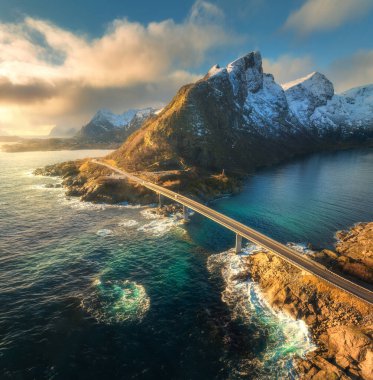 Norveç 'in Lofoten Adaları' nda günbatımında, deniz ve dalgalarla dolu bir köprü manzarası. Güzel yolu, suyu, kayaları, bulutlu mavi gökyüzünü ve altın güneş ışığını olan bir manzara. İHA 'dan üst görünüm