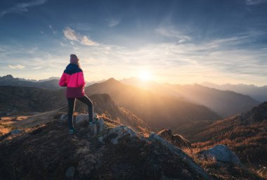 Dağın zirvesinde genç bir kadın ve sonbaharda renkli günbatımında sisli bir dağ vadisi. Dolomitler, İtalya. Sportif kız, sisli dağ tepeleri, turuncu çimenler, ağaçlar, sonbaharda altın güneş. Yürüyüş