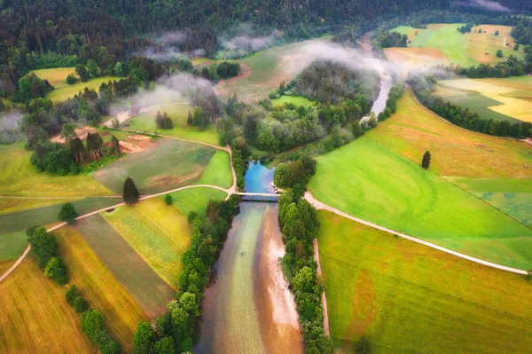 Aerial view of beautiful river in low clouds at sunrise in summer in Slovenia. Turns of river, green meadows and fields, grass and trees, rural road at dawn in spring. Top drone view of countryside
