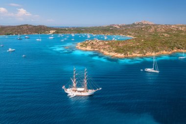 Yazın günbatımında mavi denizde yelkenli ve lüks yatların hava manzarası. Sardunya, İtalya. Sürat tekneleri, yatları, deniz koyları, dağ, açık gök mavisi suyu ve gökyüzü olan tropik deniz manzarası. Okyanus manzarası