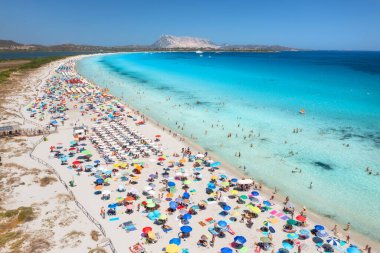 Renkli şemsiyeler, beyaz kumlu plaj, insanlar, mavi deniz, gök mavisi su, yaz güneşli bir günde dağ. Tropik deniz manzarası. İtalya, Sardunya 'da seyahat ve tatil. Deniz kenarının en üst drone görünümü