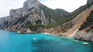 Kayaların ve dağların havadan görünüşü, deniz körfezi, sahil, yaz günü tekneler. Cala Goloritze, Sardunya Adası, İtalya. Uçurumların, taşların, yeşil ağaçların, gün batımında şeffaf masmavi suyun en üst drone görüntüsü
