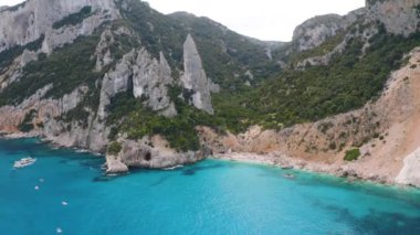 Kayaların ve dağların havadan görünüşü, deniz körfezi, sahil, yaz günü tekneler. Cala Goloritze, Sardunya Adası, İtalya. Uçurumların, taşların, yeşil ağaçların, gün batımında şeffaf masmavi suyun en üst drone görüntüsü