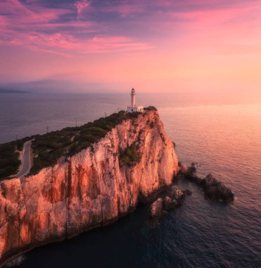 Yazın günbatımında dağın zirvesindeki deniz feneri. Hava görüntüsü. Kayalıklarda, kayalıklarda, deniz ve gökyüzünde pembe ve mor bulutlarla güzel bir deniz feneri. Lefkada Burnu, Yunanistan 'ın en iyi manzarası. İşaret
