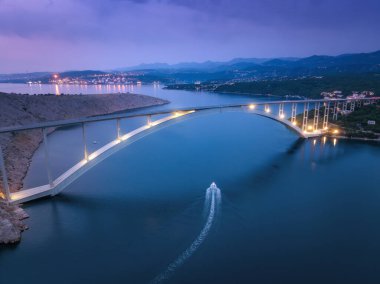 Gün batımında deniz üzerinde köprü. Aydınlatma, ada, dağlar, mavi deniz, tekne ve mor gökyüzü ile modern Krk köprüsünün havadan görünüşü. Yol manzarası, şehir ışıkları, kayalar. Mimari