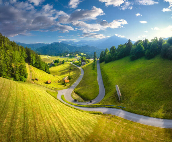 Aerial view of country road in green meadows at sunset in summer. Top drone view of rural road, alpine mountains. Colorful landscape with curved highway, hills, fields, green grass, blue sky. Slovenia