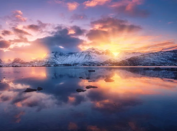 冬の日没時には 海の海岸 美しい雪の山と雲と黄金の太陽の光とカラフルな空 ノルウェーのロフテン島 雪の中の岩 夕暮れ時の水の反射 — ストック写真