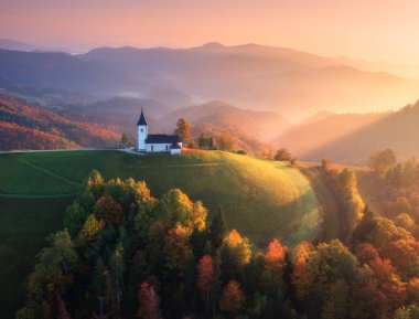 Sonbaharda gün doğumunda sisli ve renkli ormanlardaki küçük kilisenin hava manzarası. Slovenya. Dağın tepesindeki güzel şapelin sisli manzarası, yeşil çayırlar, ağaçlar, sonbaharda pembe gökyüzü. Manzara
