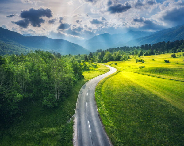 Aerial view of road in green alpine meadows at sunset in summer. Top drone view of rural road, mountains, forest. Colorful landscape with roadway, sun rays, trees, hills, green grass, clouds. Slovenia