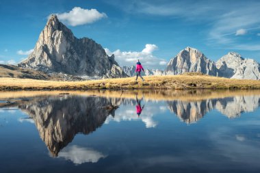 Yürüyen kadın ve dağlar, sonbaharda Dolomites, İtalya 'da günbatımında göle yansıdı. Gölün kıyısında duran kız yüksek kayalara ve sonbaharda bulutlarla kaplı mavi gökyüzüne bakıyor. Sudaki yansıma