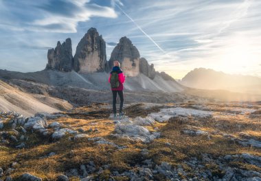 Sonbaharda gün batımında dağ zirvesinde sırt çantalı ve yüksek kayalıklı bir kız. Tre Cime, Dolomitler, İtalya. Yoldaki genç kadınla manzara, uçurumlar, bulutlu gökyüzü ve sonbaharda güneş. Yürüyüş