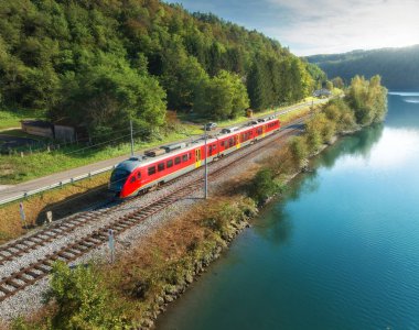 Sonbaharda günbatımında Alp Dağları 'nda nehir kıyısında hareket eden modern kırmızı hızlı trenin hava manzarası. Trenin üst manzarası, kırsal demiryolu, göl, yol, sonbaharda yeşil ağaçlar. Slovenya 'daki tren istasyonu
