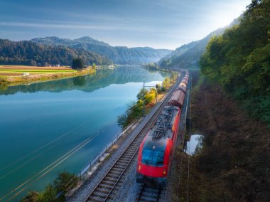 Sonbaharda güneşin doğuşunda Alp Dağları 'nda nehrin yanında ilerleyen kırmızı, modern hızlı trenin hava manzarası. Tren, demiryolu, göl, yansıma, sonbaharda ağaçlar. Slovenya 'daki tren istasyonu