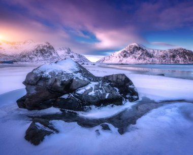 Kum kutup plajı, mavi denizi, donmuş kıyılarda taş ve kışın kar altında kayalar Lofoten Adaları, Norveç 'te günbatımında. Karlı dağları olan renkli bir manzara, bulutlu gökyüzü. Norveç kıyıları