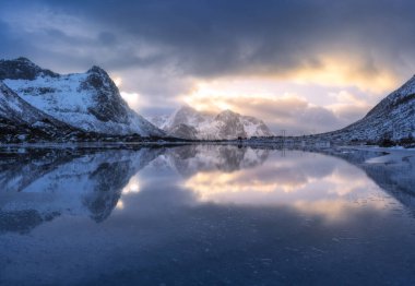 Norveç 'in Lofoten adalarında kışın gün batımında güzel karlı dağlar ve bulutlu renkli gökyüzü ve altın güneş ışığı. Karda kayalar, deniz kıyıları, suda yansıma, bulutlu gökyüzü.