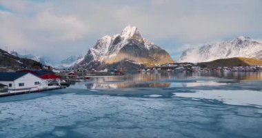 Karlı kayaların havadan görünüşü, daha karlı adalar, donmuş deniz kıyıları, dağlar, yollar, kışın gün batımında bulutlu gökyüzü. Norveç, Lofoten Adaları 'ndaki Reine köyünün güzel insansız hava aracı manzarası