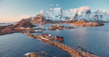 Karlı kayaların havadan görünüşü, deniz bölmesi, köprü, dağlar, yol, kışın gündoğumunda mavi gökyüzü. Norveç, Lofoten Adaları 'ndaki Hamnoy ve Reine köyünün güzel insansız hava aracı manzarası. Doğa