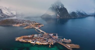 Karlı kayaların havadan görünüşü, deniz, köprü, dağlar, yol, kışın dramatik gündoğumunda bulutlu gökyüzü. Norveç, Lofoten Adaları 'ndaki Hamnoy ve Reine köyünün insansız hava aracı görüntüsü. Rorbuer