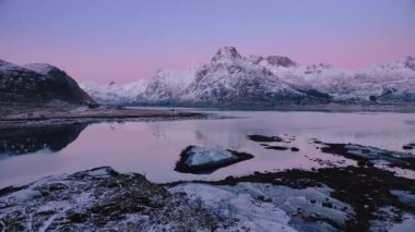 Deniz manzarası, karlı dağlar, kışın gün batımında daha kötü. Lofoten Adaları, Norveç. Fiyort, ev, sudaki yansıma, taşlar, kayalar, alacakaranlıktaki pembe bulutlu mor gökyüzü. Göl