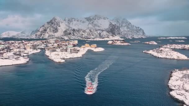 雪の降るヘニングヴァー村 ノルウェーのロフテン諸島の青い海での浮遊漁船の空中観察 ボートのトップドローンビュー 雪の山 曇った空 — ストック動画