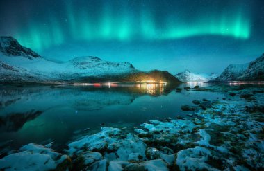 Karlı dağların üzerinde Aurora borealis, donmuş deniz, Norveç, Lofoten 'de kış gecelerinde suda yansıma. Kuzey ışıkları ve karlı kayalar. Kutup ışıkları, taşlar, yıldızlı gökyüzü ve fiyortlu manzara