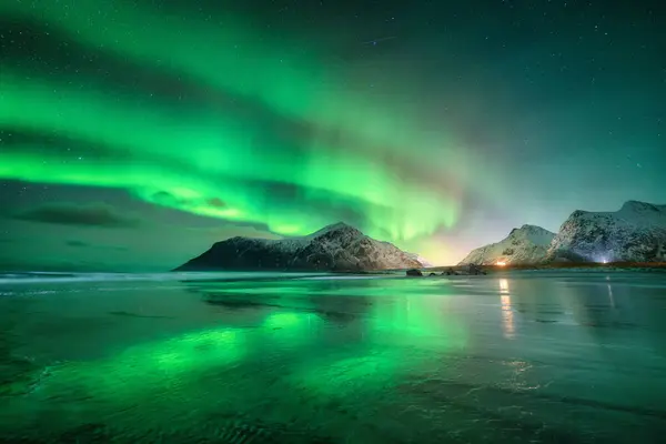 Northern Lights and sandy beach at starry winter night. Lofoten islands, Norway. Beautiful Aurora borealis. Sky with polar lights. Landscape with aurora, sea, sky reflection in water, snowy mountains