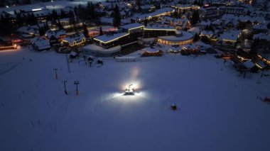 Slovenya 'nın Kranjska Gora köyünde kış gecesi karlı alp kayak yamacında çalışan kar arabasının hava görüntüsü. Kar temizleme, kayak merkezi, alacakaranlıkta sokak lambaları. Kar küreme aracı