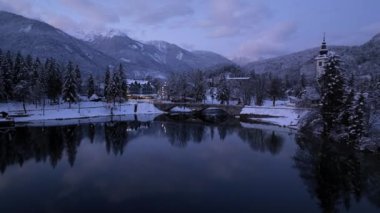Slovenya 'daki Bohinj Gölü' nün kışın alacakaranlıktaki havadan görünüşü. Karlı dağların, ağaçların, kilisenin, köprünün, yolun, şehrin ışıklarının, evlerin, sudaki yansımanın, gün batımında bulutların olduğu mor gökyüzünün tepedeki drone görüntüsü.