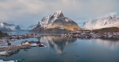 Karlı kayaların havadan görünüşü, deniz koylu adalar, dağlar, yol, kışın gün doğumunda alçak bulutlu gökyüzü. Norveç, Lofoten Adaları 'ndaki Reine köyünün güzel insansız hava aracı manzarası